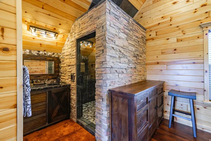 Rustic cabin bathroom featuring knotty pine wood walls and ceiling, custom stone accent wall with integrated shower enclosure, dark wood vanity with mirror, and warm wood-finish flooring throughout