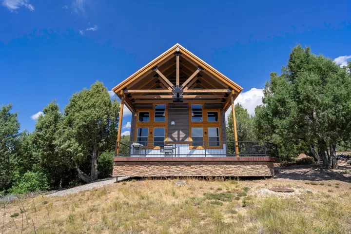 Gable roofline with exposed wood beams, wood siding exterior, stone veneer foundation, expansive deck with black metal railing, and surrounding mature trees