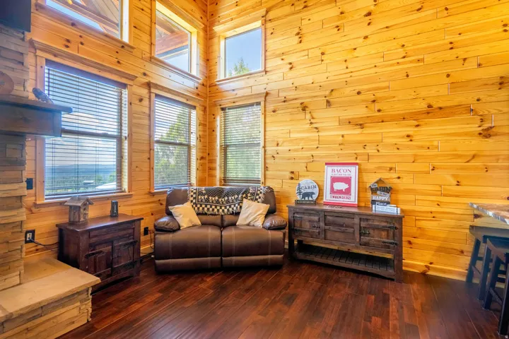 Hardwood flooring, natural wood paneling, stacked stone fireplace, multiple windows, and high ceilings