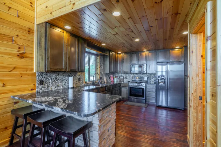 Wood-paneled ceilings, wood-finish flooring, a stone-faced breakfast bar with a dark countertop, stainless steel appliances, and a mosaic tile backsplash