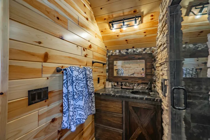 Wood plank wall and ceiling finishes, stacked stone accents, wood-finish vanity with countertop, framed mirror, and a glass-enclosed shower