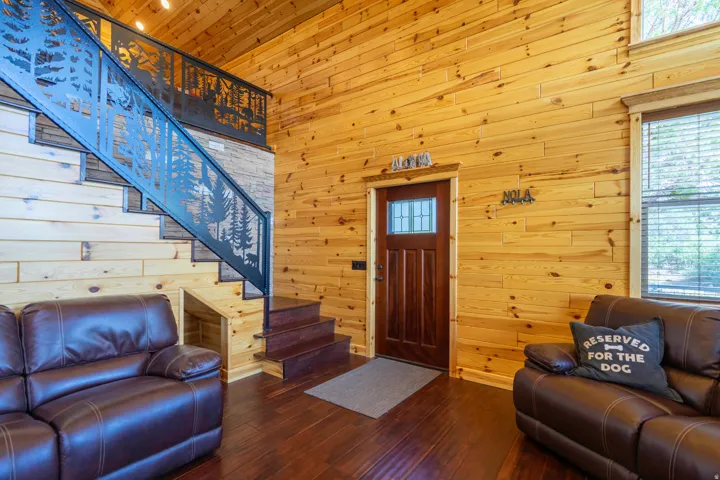 Wood-paneled interior walls and ceiling, dark wood-finish flooring, custom cut metal stair railing with forest motif, wood staircase with dark treads, and a dark wood entry door with leaded glass insert