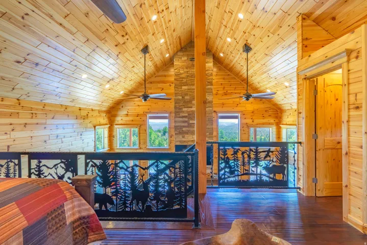 Vaulted wood-paneled ceiling with recessed lighting, dark wood-finish flooring, decorative metal railing, and a central stone-clad column