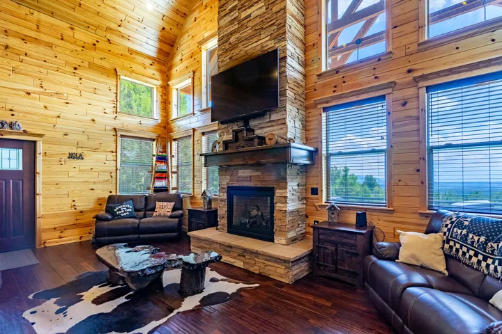Vaulted wood-paneled ceiling with exposed beams, stacked stone fireplace with wood mantel, wood-finish flooring, and multiple windows