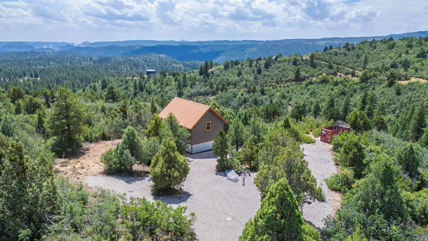 Wood-sided residence with an orange-brown metal roof, an attached garage, and a gravel driveway, surrounded by abundant evergreen trees and distant mountains