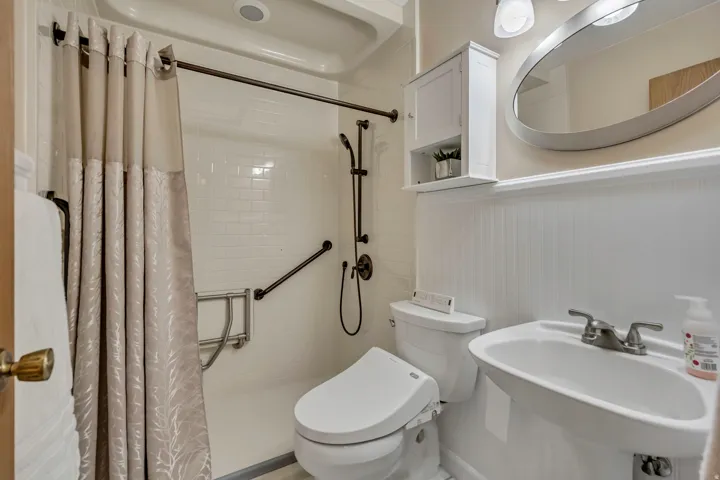 Pedestal sink with chrome-finish faucet, white beadboard wainscoting, wall-mounted storage cabinet, oval mirror, and white subway tile shower surround