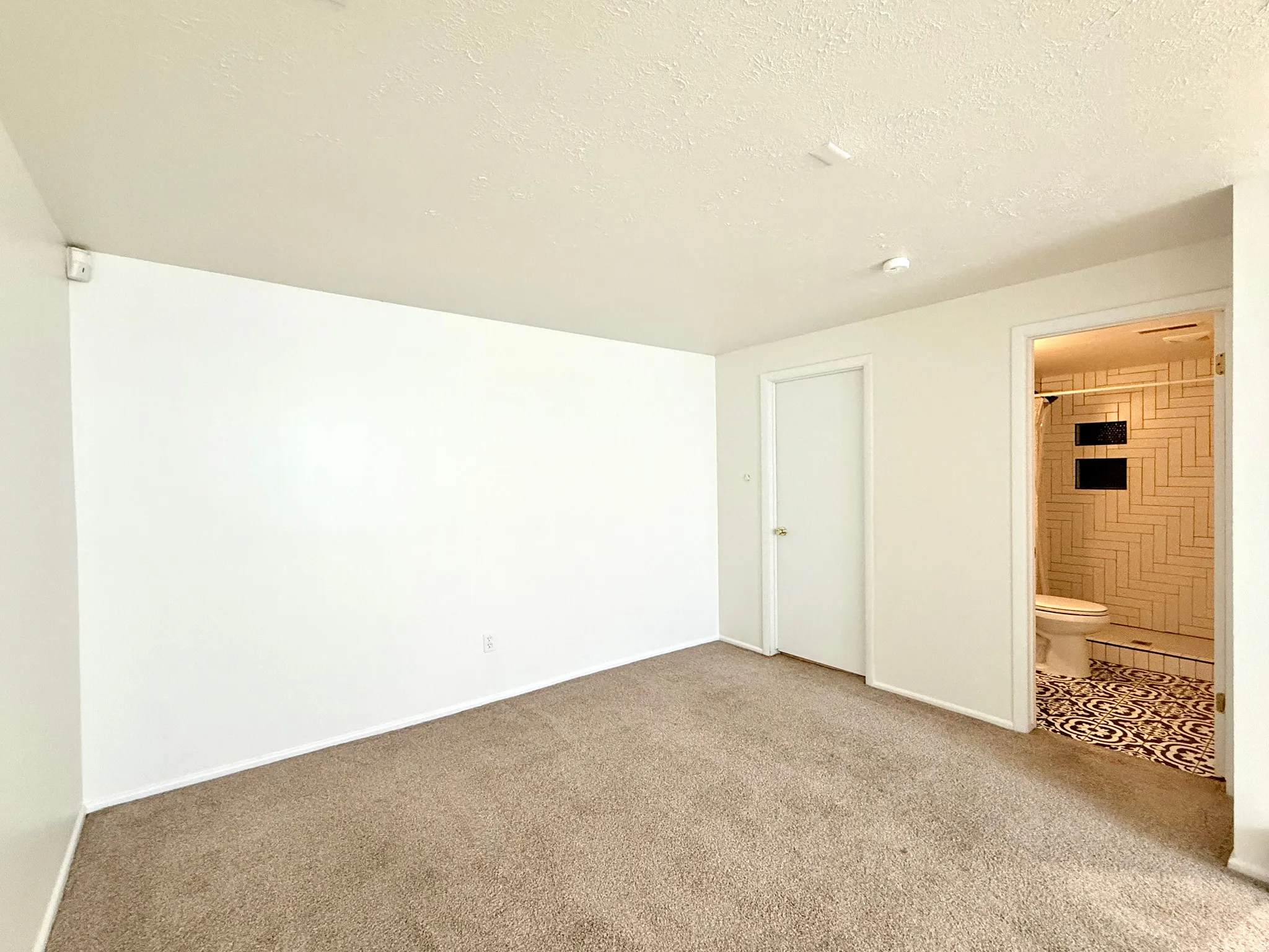 Carpeted room with recessed lighting and a white door, featuring an attached bathroom with patterned floor tiling, white subway tile shower, and built-in shower niches