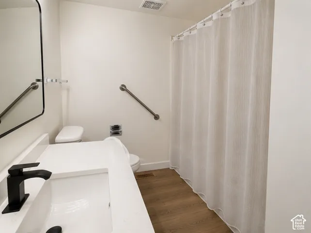 Bathroom featuring a vanity with a white countertop and rectangular undermount sink, matte black faucet, wall-mounted mirror, wood-finish flooring, and metal safety grab bars