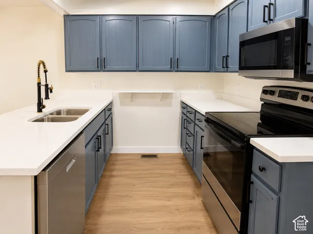 Kitchen featuring slate-blue cabinetry with matte black hardware, white solid-surface countertops, wood-finish flooring, stainless steel appliances, and a high-arc pull-down faucet