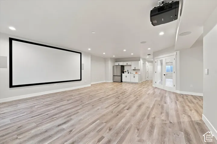 Large cozy entertainment space featuring a large projection screen, mounted projector, recessed lighting, wood-finish flooring, and a compact kitchenette with white cabinetry.