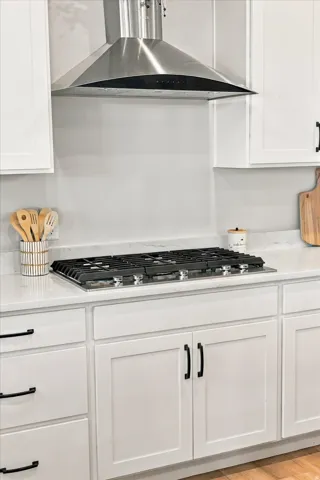 Stainless steel range hood, built-in gas cooktop, quartz-finish countertops, white shaker cabinetry, and matte black hardware.