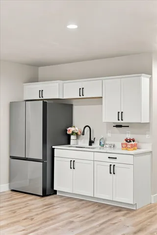 Kitchenette featuring white shaker-style cabinetry with matte black hardware, a stainless steel refrigerator, white countertops, and wood-finish flooring.