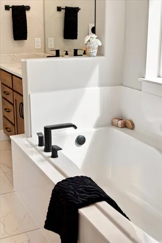 White soaking tub with matte black fixtures, adjacent to a wood-finish vanity with a light-colored countertop