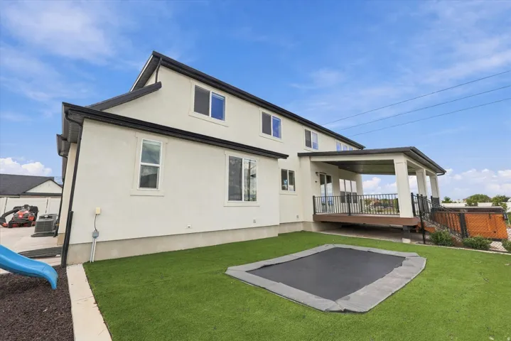 Rear exterior featuring a covered deck with railing, stucco siding, and  backyard with artificial turf and live grass areas.