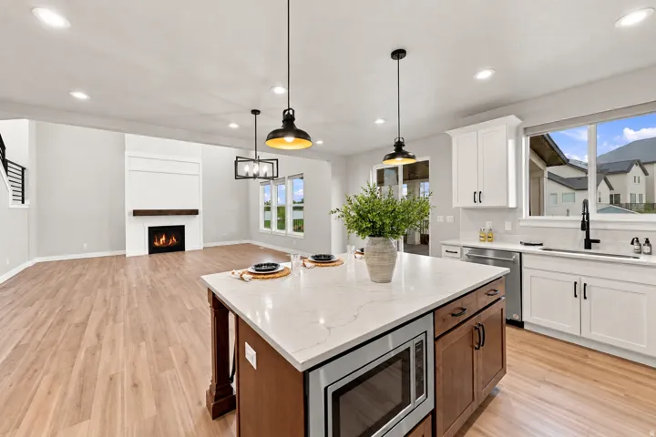 Open-concept living space featuring wood-finish flooring, a contemporary fireplace with a wood mantle, white cabinetry, a large kitchen island with a built-in microwave, and pendant lighting with lots of windows.