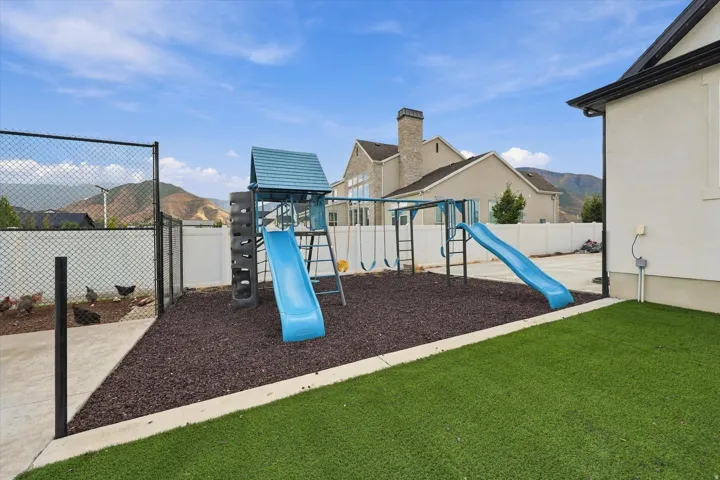 Artificial turf lawn, built-in play structure with slides and swings, chain link fencing, white privacy fencing, and concrete patio area.