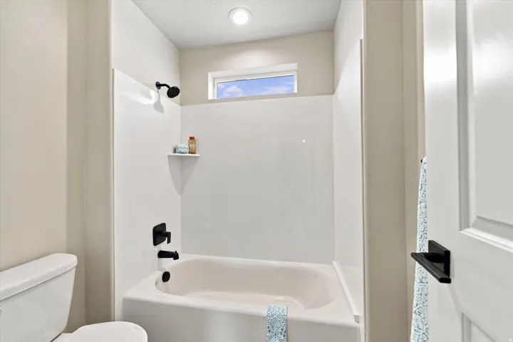 White bathtub and shower combination with matte black fixtures, an integrated shelf, and an overhead window.
