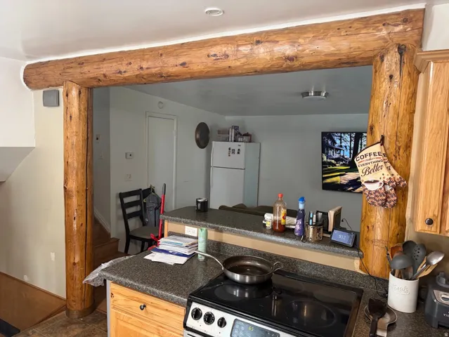 Kitchen with a built-in electric range, granite-look countertop, wood-finish cabinetry, and a pass-through opening featuring exposed log beams
