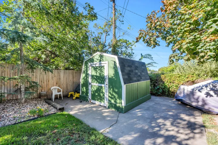 Detached storage shed with a gambrel roof and asphalt shingles, concrete pathway, fenced perimeter, and established trees