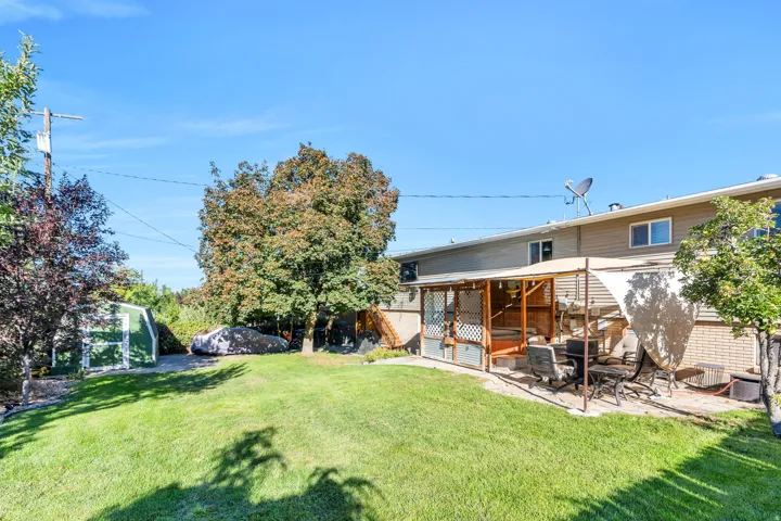Expansive backyard featuring a green lawn, mature trees, a storage shed, and a covered patio area with a stone paver surface