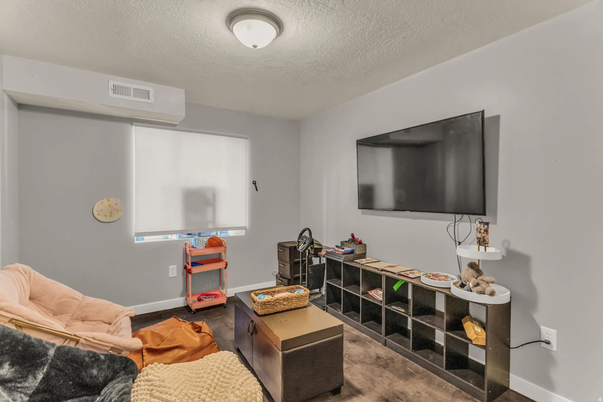 Recessed ceiling light fixture, wall-mounted television, wood-finish flooring, and light-filtering window shade