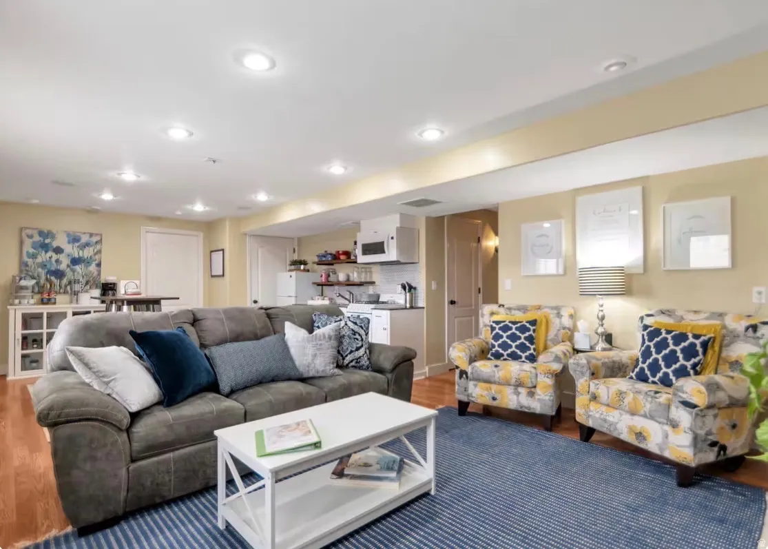 Recessed lighting illuminates this open living area featuring wood-finish flooring and a built-in kitchenette with white cabinetry and subway tile backsplash
