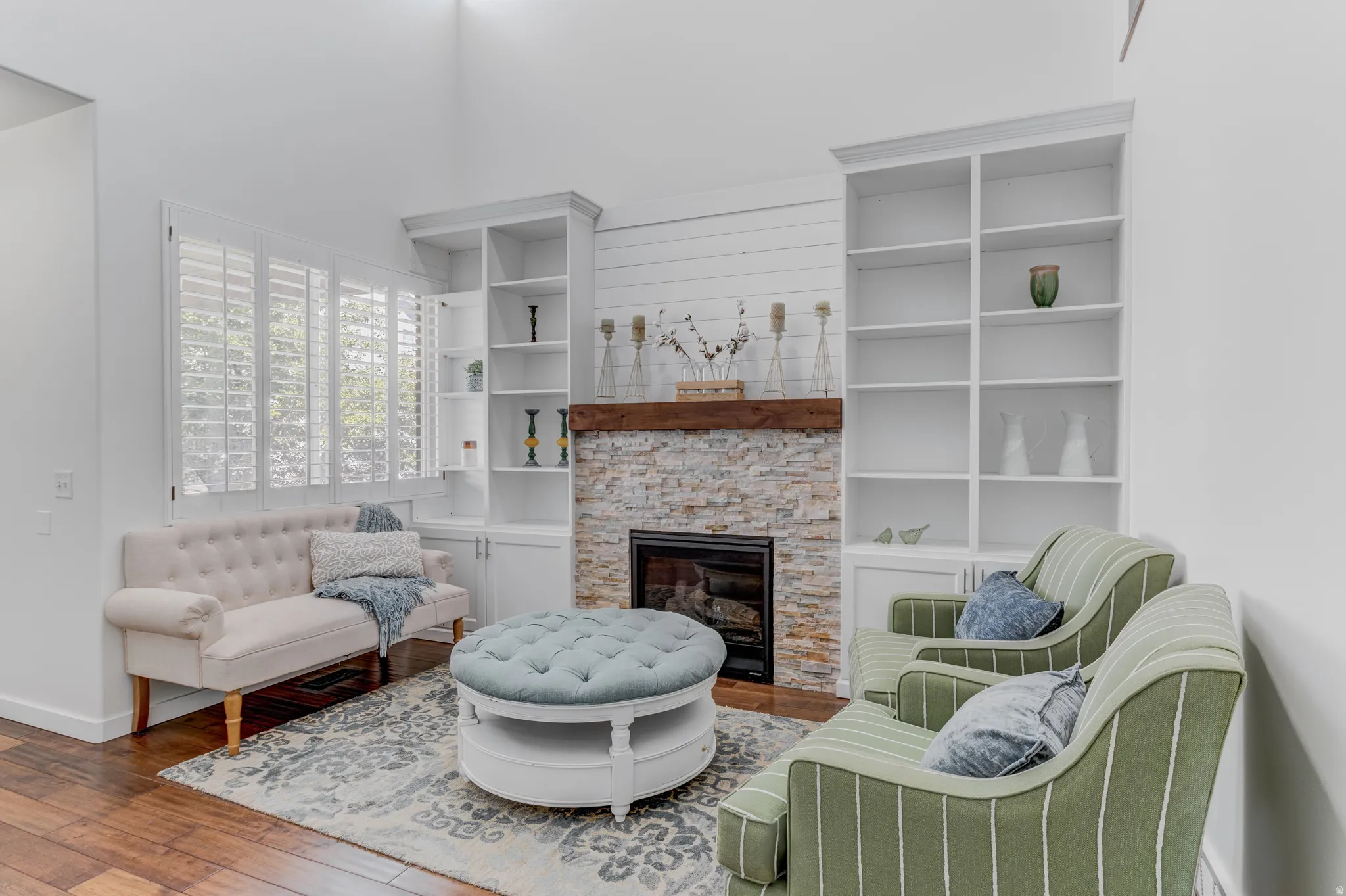 Stone-clad fireplace with wood mantel, flanked by built-in shelving, wood-finish flooring, and white plantation shutters