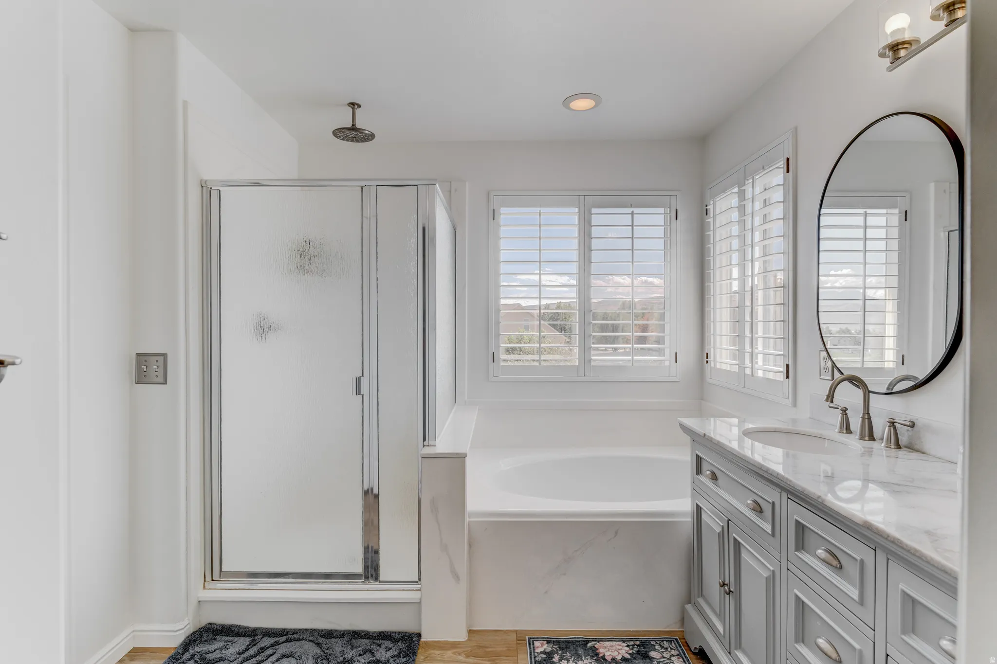 Bathroom featuring a built-in soaking tub, a separate glass-enclosed shower with a rainfall showerhead, a vanity with a stone countertop and an undermount sink, and plantation shutters