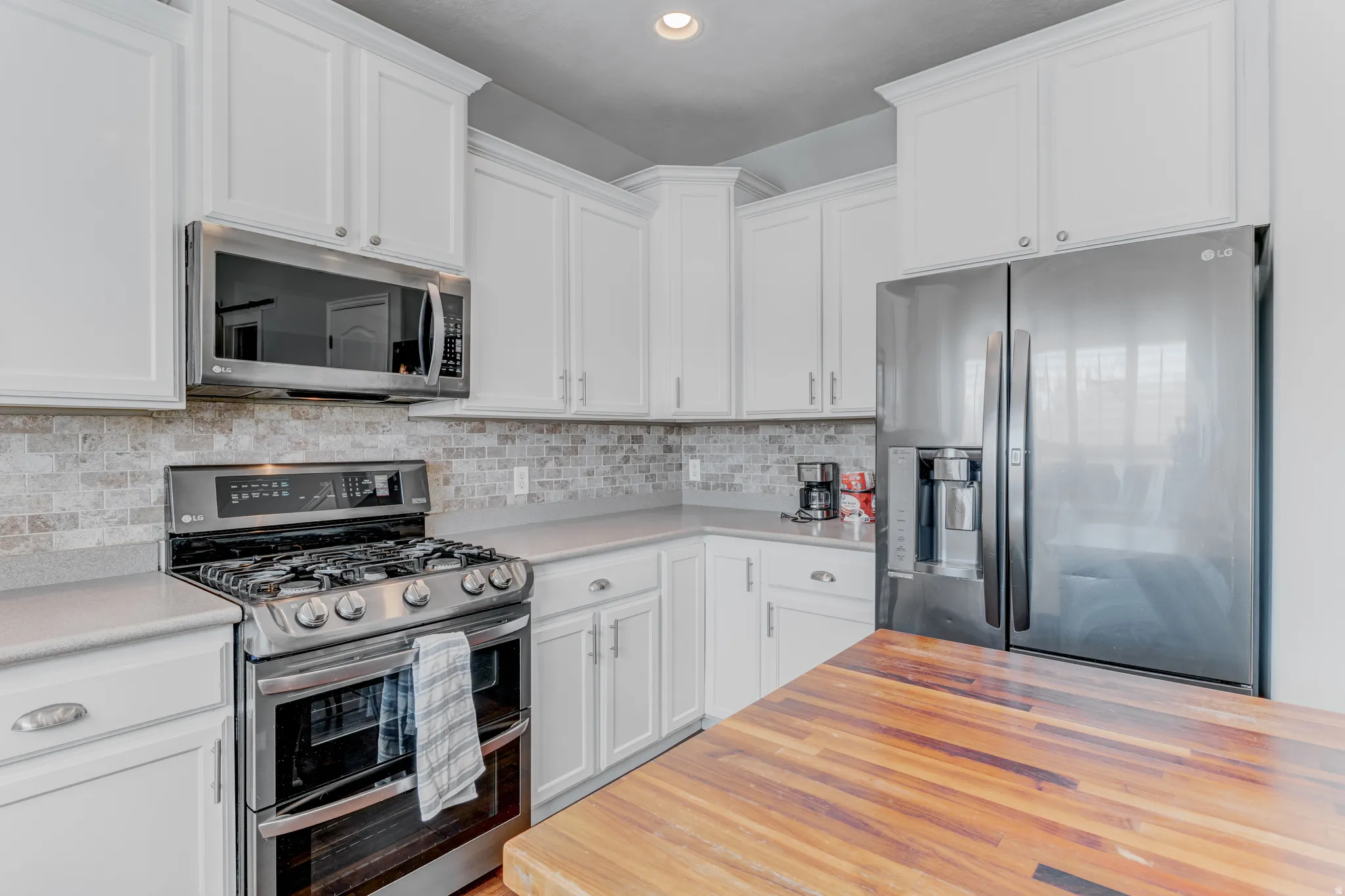 White cabinetry with brushed nickel hardware, stainless steel appliances, light-toned countertops, tile backsplash, and a wood-finish surface