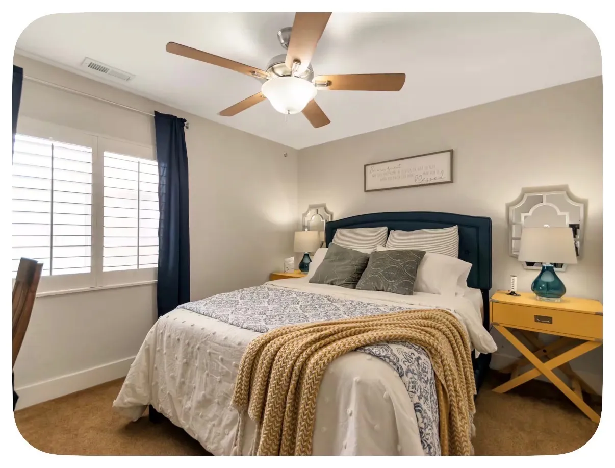 Ceiling fan with light fixture, window with white plantation shutters, upholstered headboard, and carpeted flooring