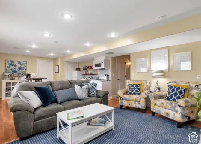 Recessed lighting illuminates this open living area featuring wood-finish flooring and a built-in kitchenette with white cabinetry and subway tile backsplash
