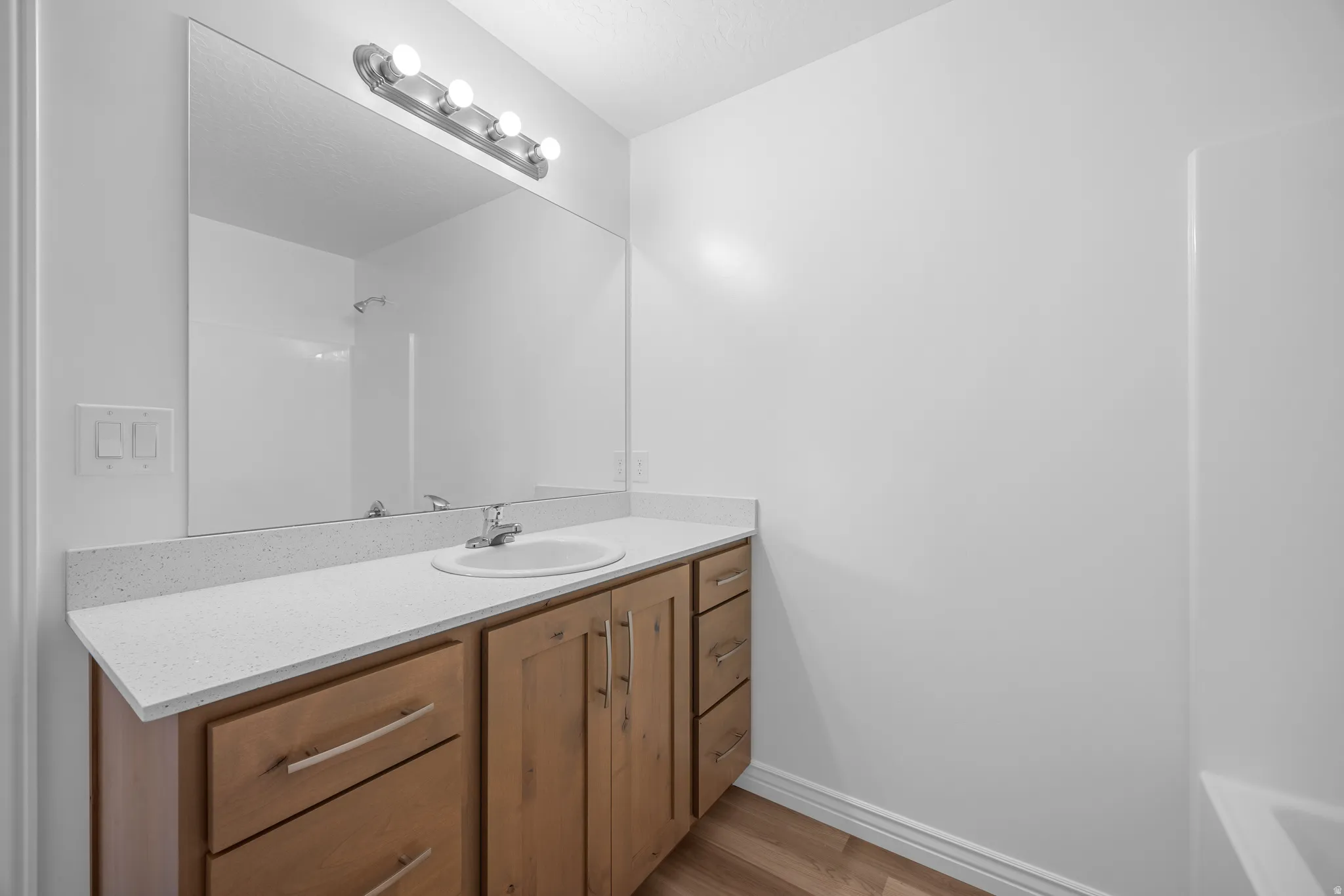 Wood-finish flooring, light wood cabinetry, white stone-finish countertop, single basin sink, and a large wall-mounted mirror