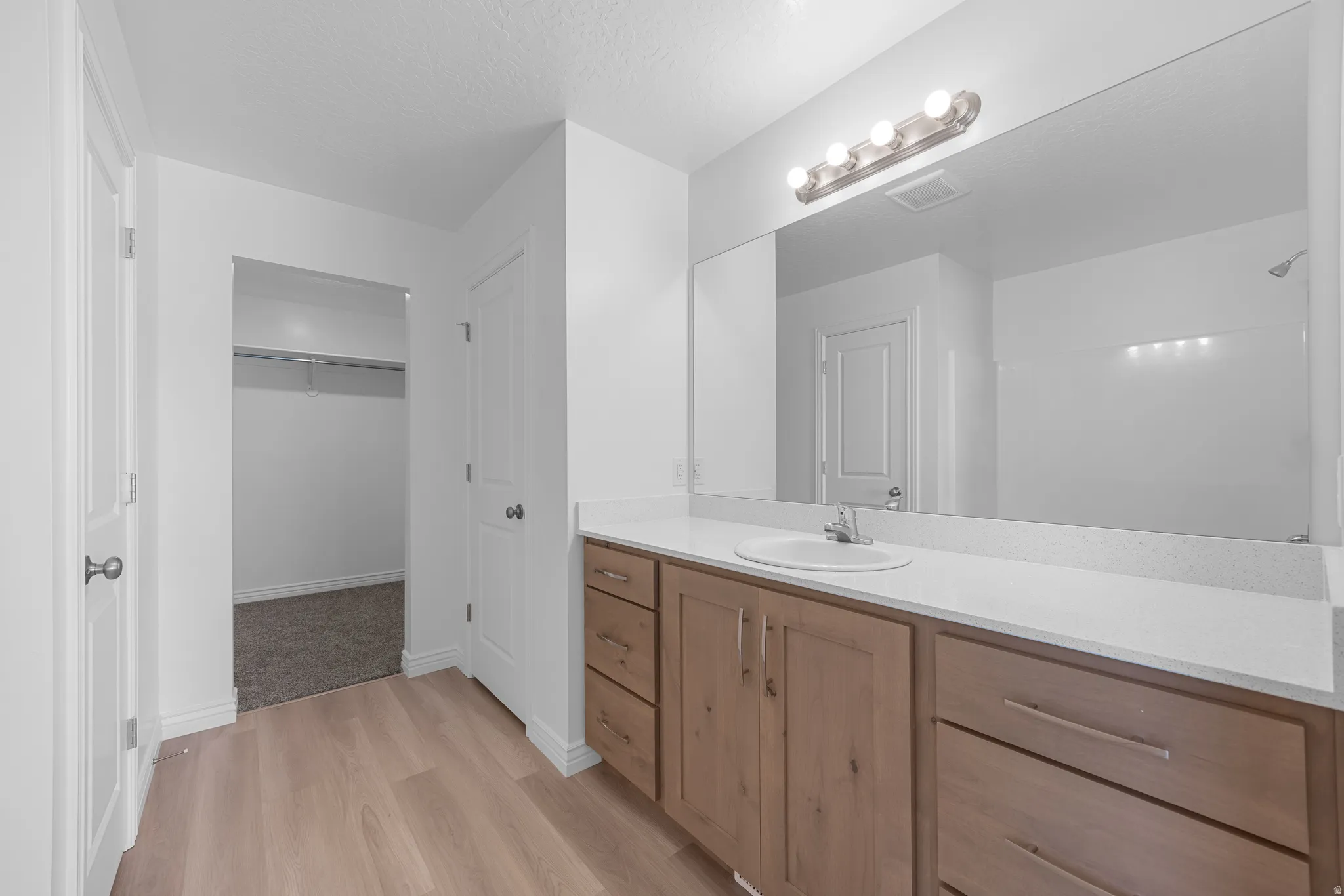 Wood-finish flooring, white solid-surface countertop, wood-finish vanity with ample storage, large vanity mirror, and modern vanity lighting