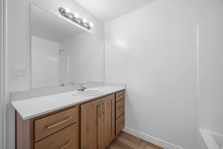 Wood-finish flooring, light wood cabinetry, white stone-finish countertop, single basin sink, and a large wall-mounted mirror