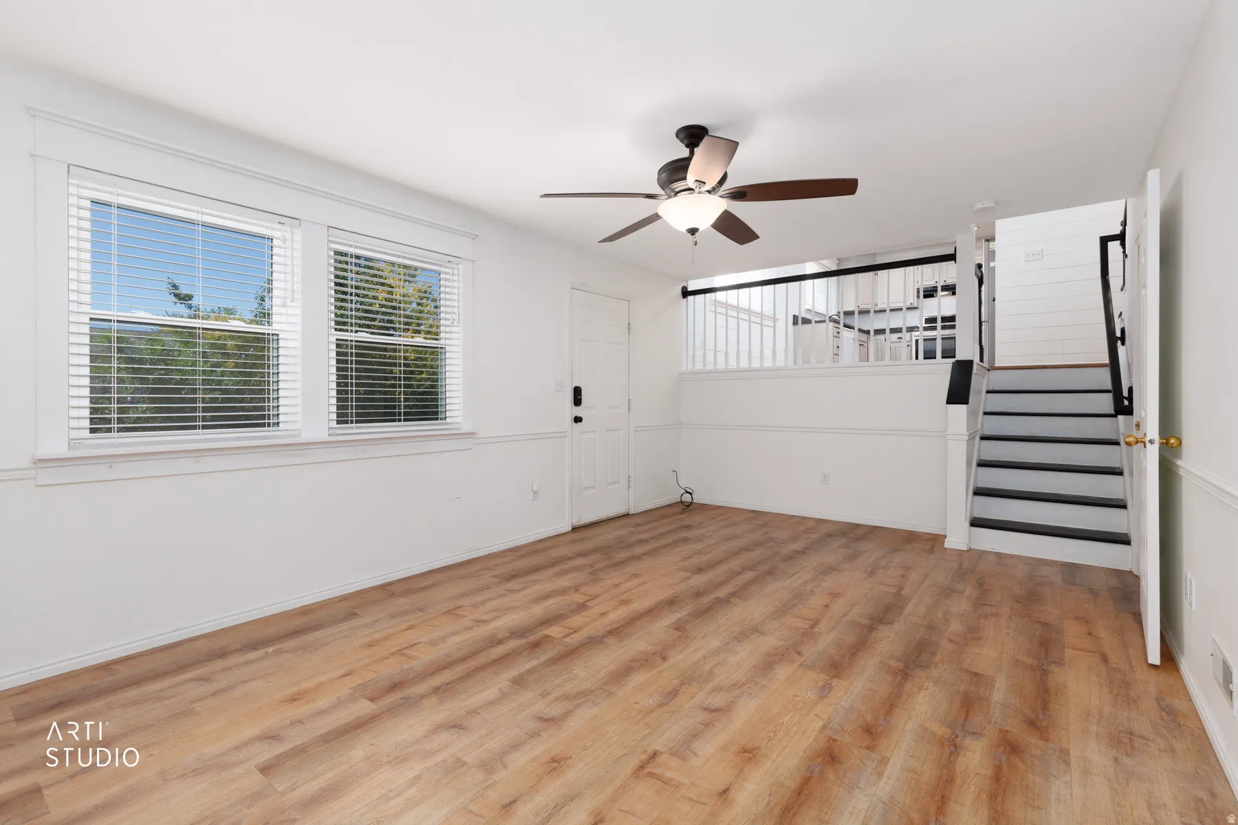 Wood-finish flooring, two large windows with blinds, ceiling fan with light fixture, paneled walls, and staircase with black handrails