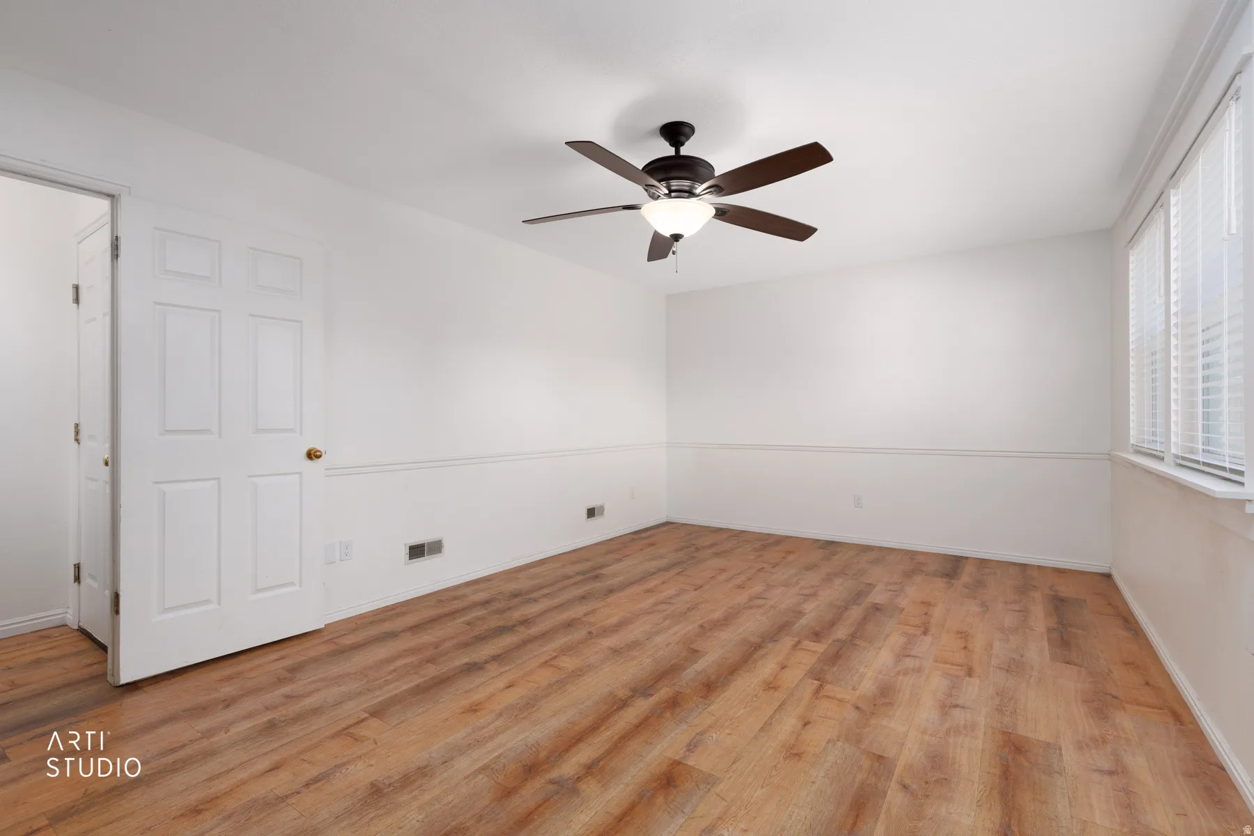 Wood-finish flooring, white walls with wainscoting, multi-pane window with blinds, ceiling fan with integrated lighting, and a six-panel door with brass hardware