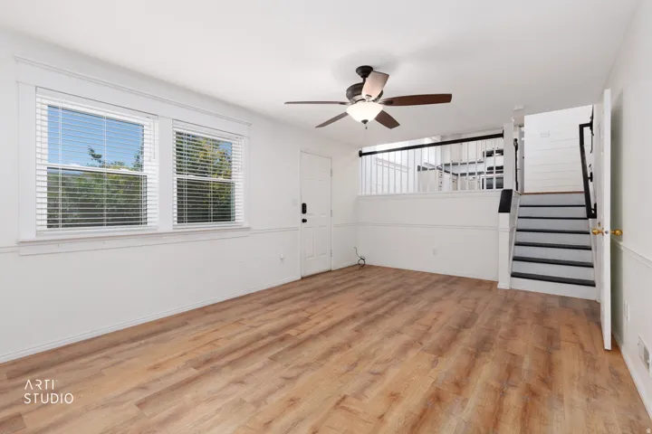 Wood-finish flooring, two large windows with blinds, ceiling fan with light fixture, paneled walls, and staircase with black handrails