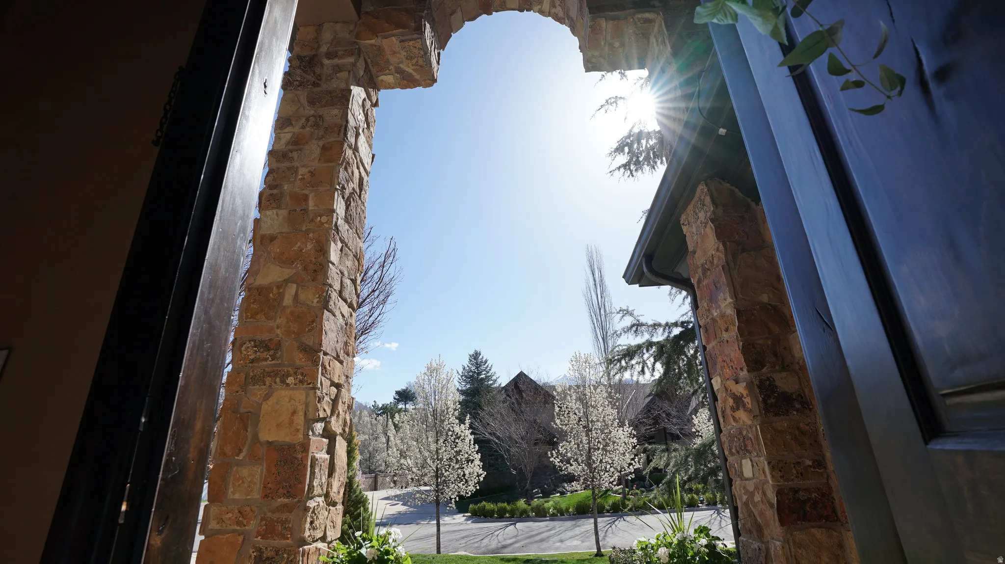 Archway entry with stone masonry, dark wood door, lush landscaping, and flowering trees