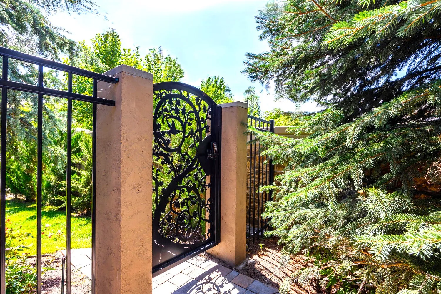 Wrought iron gate with ornate detailing, flanked by stucco-finished pillars and a matching wrought iron fence