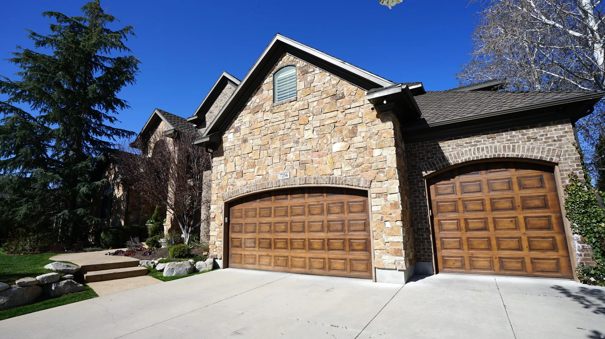 Stone and brick exterior with arched wood-finish garage doors, concrete driveway, and mature landscaping