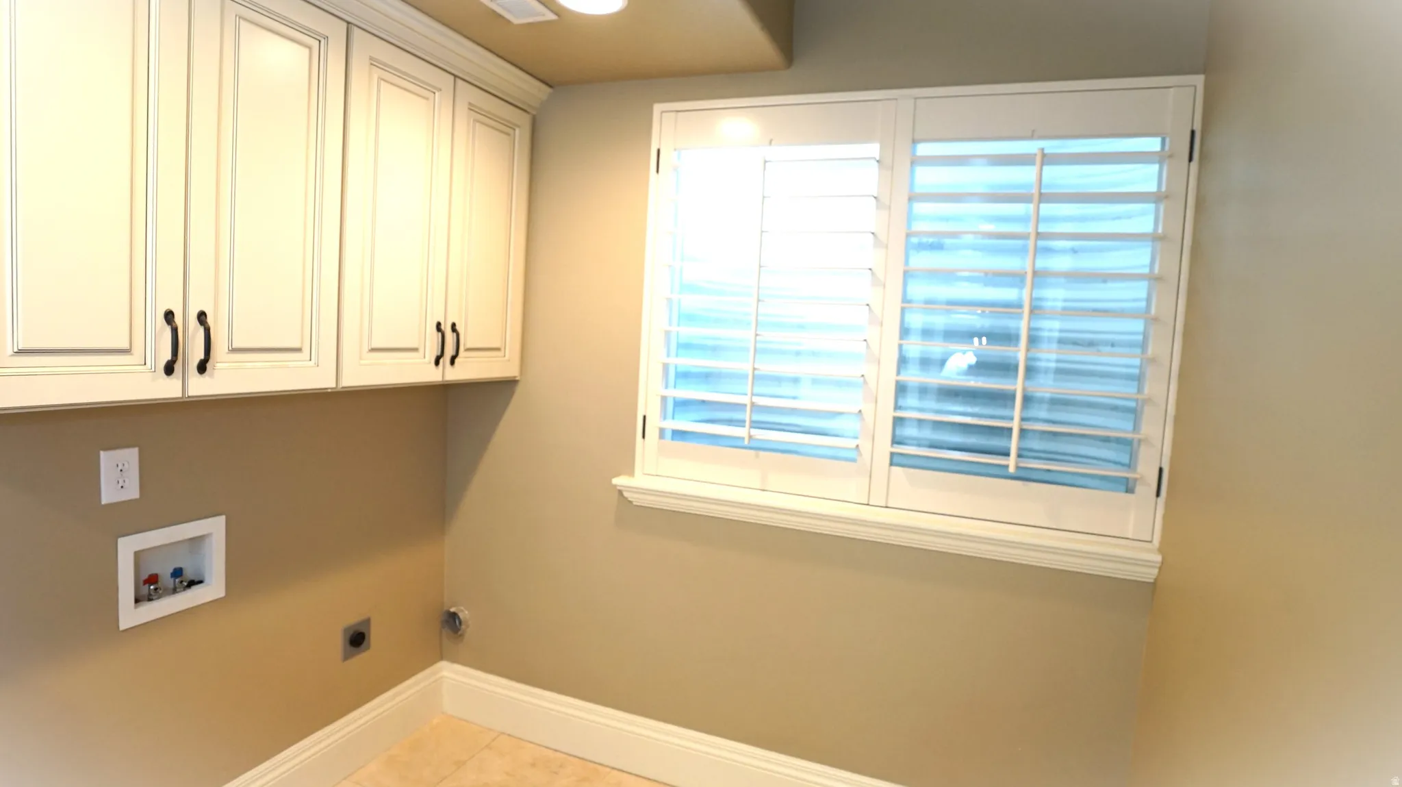 Cream-painted cabinetry with black hardware, window with white plantation shutters, and light-toned tile flooring