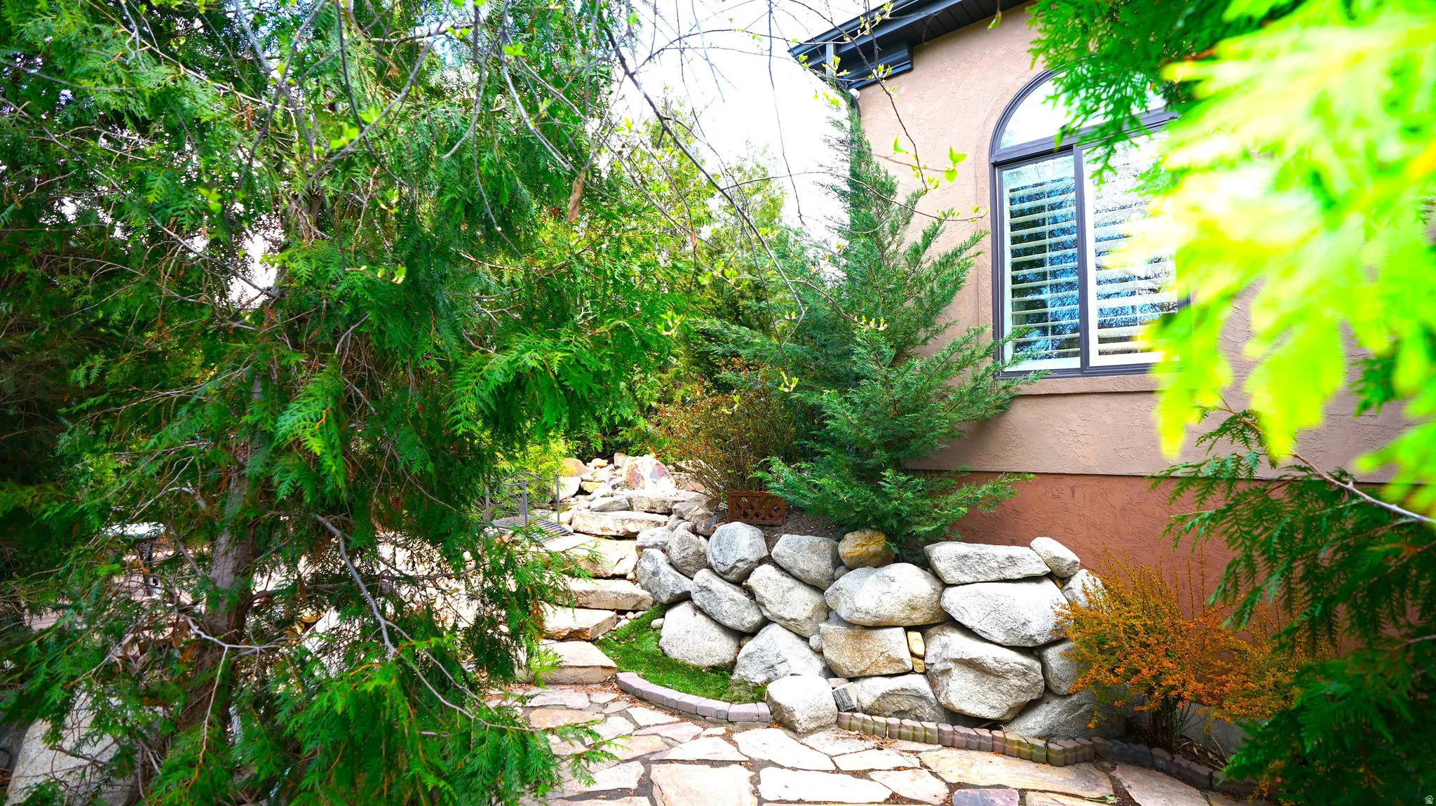 Stone pathway with natural stone steps and retaining wall, complemented by mature evergreen landscaping and a stucco exterior with an arched window
