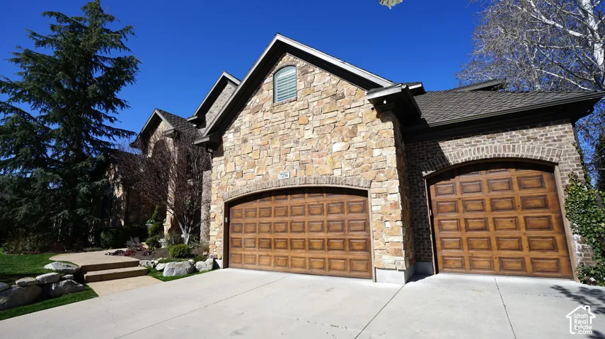 Stone and brick exterior with arched wood-finish garage doors, concrete driveway, and mature landscaping