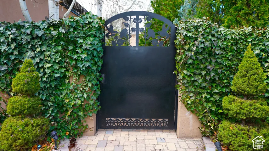 Wrought iron gate with decorative elements, flanked by ivy-covered walls, mature shrubbery, and paver stone walkway