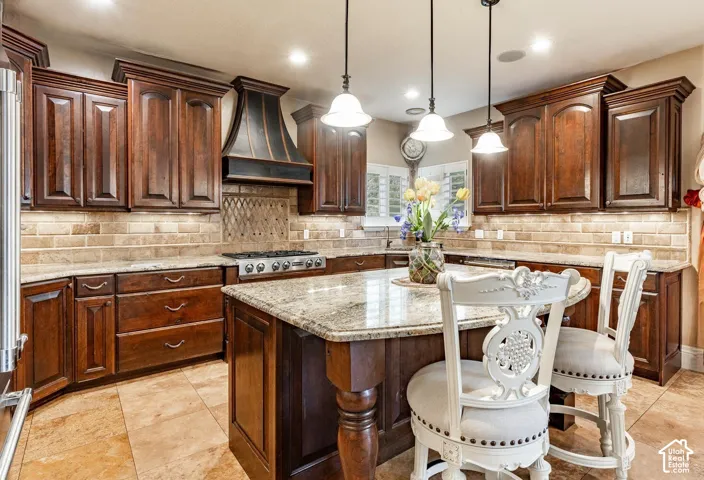 Gourmet kitchen featuring rich wood cabinetry, granite countertops, a built-in gas range with a custom range hood, a spacious central island, and stone tile flooring
