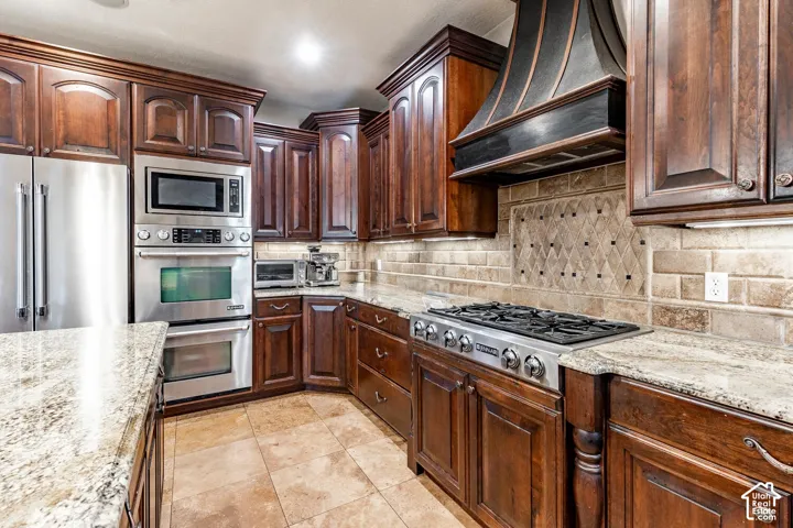 Dark wood cabinetry, stainless steel appliances, a built-in oven and microwave, stone countertops, and a decorative tile backsplash