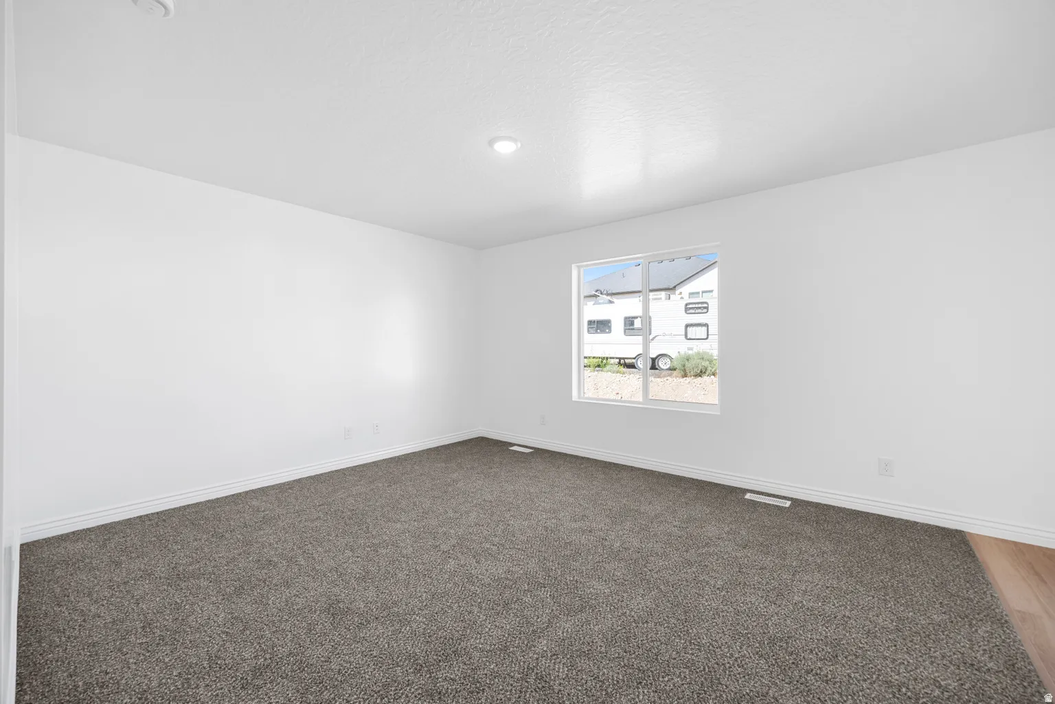Recessed ceiling light, white painted walls, gray carpeting, wood-finish flooring, and a single window with white trim
