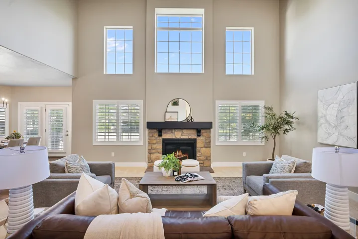 Two-story living area featuring a stone-faced fireplace with a dark wood mantel, high ceilings, multiple transom windows, and plantation shutters