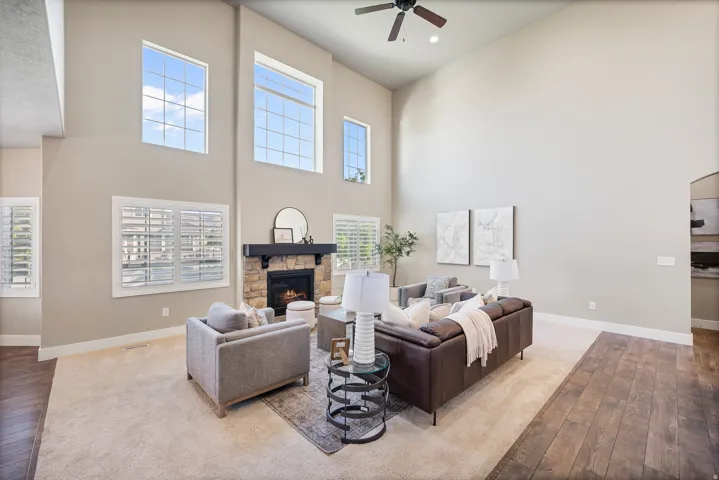 Vaulted ceiling, stone-clad fireplace with dark wood mantel, multiple windows with grid patterns, ceiling fan, and carpeted flooring