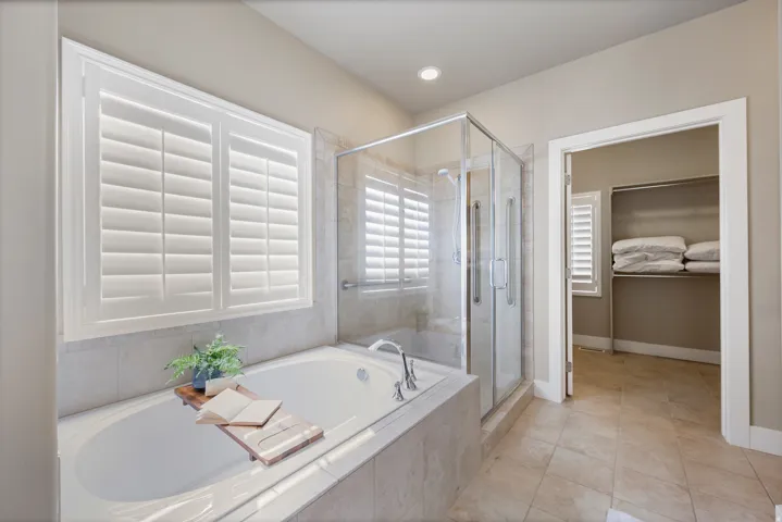 Built-in soaking tub with tile surround, glass-enclosed shower, white plantation shutters, and a walk-in closet with built-in shelving